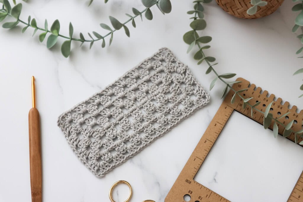 A crocheted granny rectangle swatch in light grey yarn displayed on a white surface with a wooden crochet hook, scissors, and a measuring frame surrounded by greenery.