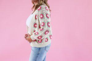 A woman wearing a handmade crochet cardigan with colorful granny square motifs in pink, green, and cream, styled with a white top and light blue jeans against a pink background.
