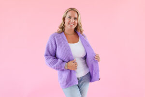 Woman wearing a handmade lilac crochet cardigan, styled with a white top and jeans, standing against a pink background showcasing the finished crochet cardigan pattern