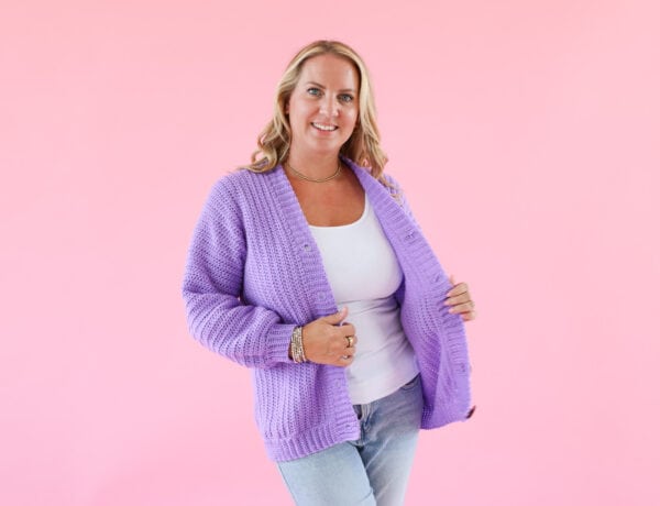 Woman wearing a handmade lilac crochet cardigan, styled with a white top and jeans, standing against a pink background showcasing the finished crochet cardigan pattern