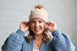 A woman wearing a beige crocheted beanie with a faux-fur pom-pom.