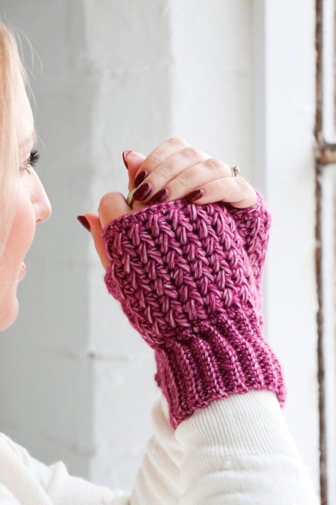 Hands wearing textured crochet wrist warmers in a deep pink yarn, shown against a white brick wall. The wrist warmers feature ribbed cuffs and a raised, textured stitch pattern.