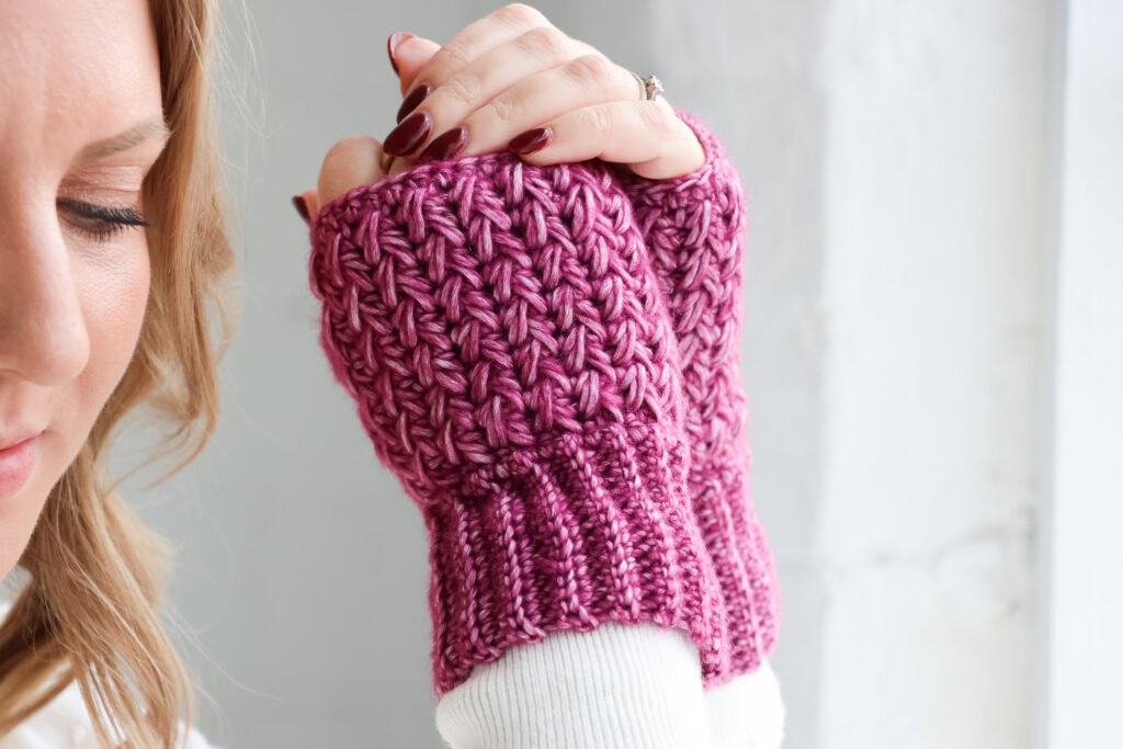Hands wearing textured crochet wrist warmers in a deep pink yarn, shown against a white brick wall. The wrist warmers feature ribbed cuffs and a raised, textured stitch pattern.