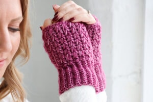 Hands wearing textured crochet wrist warmers in a deep pink yarn, shown against a white brick wall. The wrist warmers feature ribbed cuffs and a raised, textured stitch pattern.