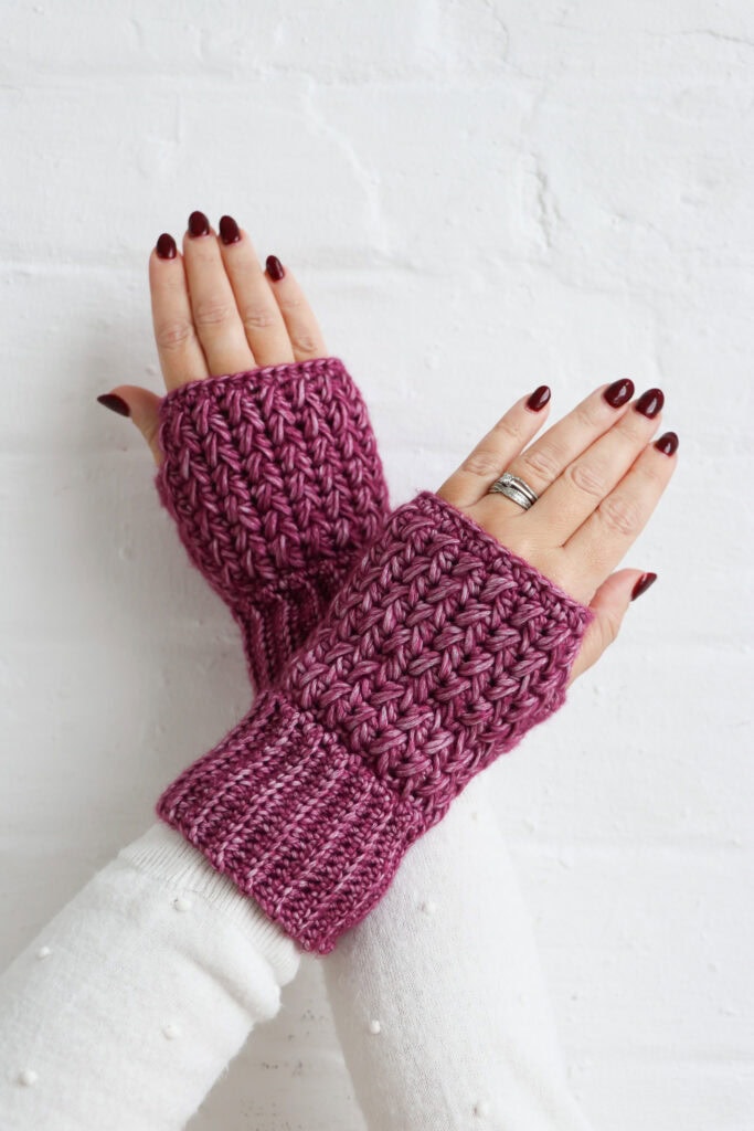 Hands wearing textured crochet wrist warmers in a deep pink yarn, shown against a white brick wall. The wrist warmers feature ribbed cuffs and a raised, textured stitch pattern.
