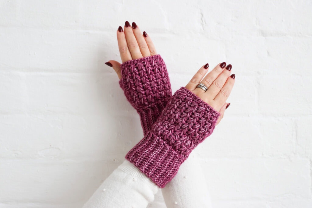Hands wearing textured crochet wrist warmers in a deep pink yarn, shown against a white brick wall. The wrist warmers feature ribbed cuffs and a raised, textured stitch pattern.